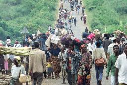 Une grande foule de personnes de tous âges portant de la nourriture et des effets personnels se dirige vers la caméra sur un long chemin de terre traversant un paysage vert vif d’herbe et de buissons. La route et la foule s’étendent dans le lointain.