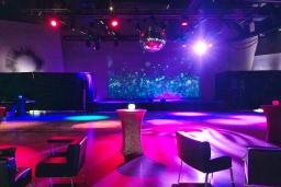 Un espace événementiel vide et coloré avec des tables de cocktail, des chaises longues et une boule disco. L'éclairage crée une atmosphère vibrante, prête à accueillir une fête ou un rassemblement.