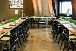 Un bistro à aire ouverte avec de longues tables en bois disposées en U et décorées de feuillage vert, d’un chemin de table blanc et d’assiettes dorées, et des chaises en cuir brun et en bois.