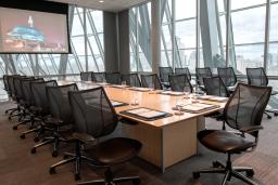 Une salle de réunion entourée de panneaux de verre et équipée d’un grand écran, d’une longue table en bois avec des cahiers et des tasses, et de chaises noires en cuir et en maille.