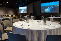 Une grande salle de classe aménagée pour une conférence ou une réunion avec deux grands écrans au mur, des tables rondes recouvertes de nappes blanches, des blocs-notes et des stylos, et des chaises en plastique gris.