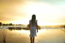 Une jeune fille aux longs cheveux noirs, vêtue d'une robe blanche jusqu'aux genoux, regarde au loin, debout jusqu'aux chevilles dans un lac calme, le soleil couchant jetant une chaude lueur jaune sur l'eau.