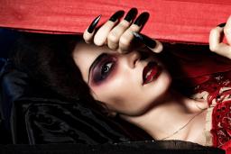 Une vampire vêtue d’une robe rouge en velours regarde à travers une boîte en bois rouge qu’elle entrouvre avec ses longs ongles noirs.