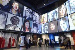 La galerie Les parcours Canadiens, du point de vue d'un visiteur debout au milieu de la galerie, entourée de niches qui racontent les expériences des Canadiens en matière de droits de la personne. Sur les murs au-dessus des niches, de grandes images de visages de défenseurs canadiens des droits de la personne sont disposées en quadrillage.