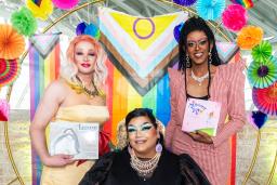 Trois artistes de drag tenant des livres se tiennent devant une arche en forme de cercle aux couleurs de l’arc-en-ciel.