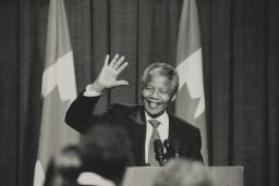 Une photographie en noir et blanc de Nelson Mandela debout derrière un podium. Il sourit et fait signe de la main.