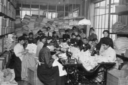 Un grand groupe de femmes assises en train de coudre. D’autres sont debout derrière elles dans une salle remplie de tissus.