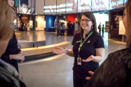 Une guide interprète debout, les bras tendus, dans une galerie de Musée. On voit des participants et participantes à la visite guidée à l'avant-plan.
