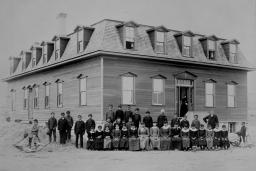 Sur cette photo en noir et blanc, un groupe d’enfants autochtones et de responsables scolaires, portant des costumes ou des robes sombres, posent devant un pensionnat.