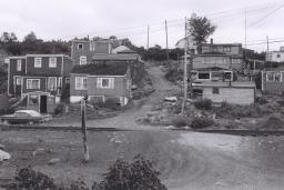 Une image en noir et blanc d’une douzaine de maisons sur une colline. Au milieu de l’image, on voit un chemin de terre qui monte la colline entre les maisons.