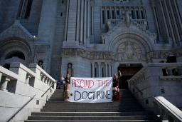 Sur les marches d’une énorme cathédrale, deux personnes portant des tresses et des jupes en ruban lèvent le poing et tiennent une grande bannière en tissu sur laquelle on peut lire en anglais « ABOLIR LA DOCTRINE ». 