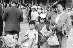 Photo en noir et blanc d’une femme avec deux enfants qui se tiennent derrière une pile de bagages et de couvertures et qui fixent l’objectif.