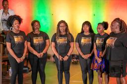Un groupe de six artistes noires debout sur scène avec un rétroéclairage coloré.