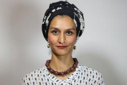 Une femme pakistanaise, Anna Abbas, portant un bandeau noir et blanc, un rouge à lèvres rouge, un collier brun épais et un chemisier noir et blanc regarde vers l'avant d'un air perplexe.