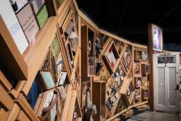 Une grande installation artistique horizontale avec un cadre en bois composé de multiples losanges et rectangles sur lequel sont fixés de nombreux objets. Une porte à moitié ouverte se trouve au milieu de l’installation.