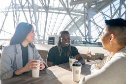 Trois personnes sont assises autour d'une table, souriantes, chacune avec une tasse à café en papier devant elle. Le soleil brille derrière elles, à travers les vitres qui entourent le Musée canadien pour les droits de la personne.