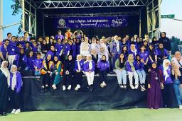Des dizaines de personnes sont rassemblées sur une scène, la plupart souriantes et vêtues de violet. Beaucoup portent le hidjab. Une banderole est suspendue à l’arrière de la scène sur laquelle on peut lire, en anglais « Prenons l’initiative. Mettons fin à l’islamophobie. #OurLondonFamily ».