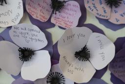 Des fleurs à trois pétales en papier de différentes nuances de violet sont regroupées dans une œuvre murale. Des noms et des messages y sont inscrits.