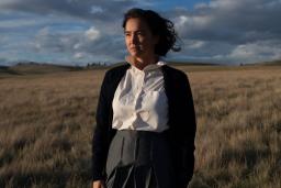 Une femme autochtone se tient dans un champ d’herbes de prairie. Derrière elle, le ciel bleu est parsemé de nuages gris et le soleil l’éclaire.