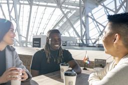 Trois personnes sont assises à une table. Elles sourient toutes, un café devant elles. Un petit drapeau de la Fierté progressif est posé sur leur table et sur la table vide derrière elles. Le soleil brille à travers les fenêtres du Musée.