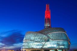 Un grand édifice avec une façade en verre incurvée et une tour en verre illuminée en rouge dans le crépuscule.