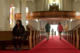 Intérieur d’une église avec une longue allée recouverte d’un tapis rouge entre des bancs en bois. La lumière du soleil filtre à travers les vitraux tandis que plusieurs enfants courent vers l’entrée lumineuse au fond de l’église et qu’une femme autochtone est assise seule sur un banc à gauche.