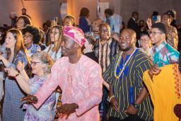 Un groupe de personnes de tous âges, dont beaucoup portent des vêtements africains traditionnels aux couleurs vives, dansent et sourient.