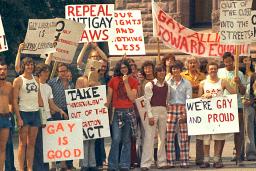 Un grand groupe de personnes vêtues de t-shirts et de pantalons évasés à la mode des années 1970 sont debout ensemble et tiennent des pancartes portant des messages en anglais tels que « sortis du placard », « c’est bien d’être gai » et « abrogez les lois anti-gaies ».