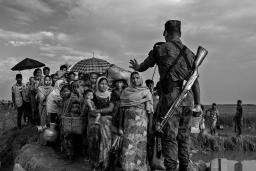 Un soldat avec un fusil en bandoulière sur le dos empêche un groupe de Rohingyas avant qu’ils ne traversent un pont de fortune. Sa main gauche est levée et fait un geste pour qu'ils s'arrêtent.