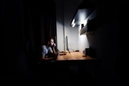 Un homme blanc aux cheveux courts est assis seul à un bureau illuminé dans une pièce sombre. Il fixe un écran d’ordinateur.
