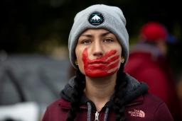 Une jeune femme aux cheveux tressés, vêtue d’une veste bordeaux et coiffée d’un bonnet d’hiver gris, regarde résolument vers l’avant, une empreinte de main rouge peinte sur la bouche et le visage.