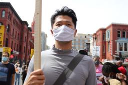 Un homme portant un masque en tissu et tenant une pancarte, marchant avec un groupe de personnes portant des pancartes sur la fin de la violence raciste et du racisme anti-asiatique.