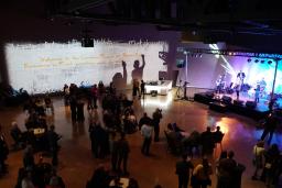 A crowded hall of people watching musicians perform on a brightly lit stage. On the wall in the background, a projected image reads “Welcome to the Canadian Museum for Human Rights.” 