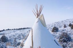 Un tipi recouvert de neige se dresse dans un paysage vallonné parsemé d’arbres et de buissons, sous un ciel bleu.