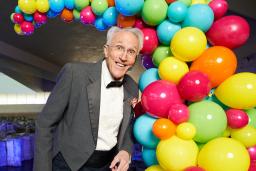 Un artiste portant un complet se tient devant des ballons multicolores. 