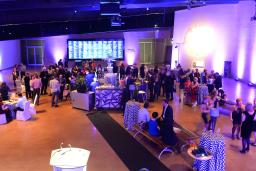 Une fête se déroule dans une grande salle. Les gens sont rassemblés autour de tables et se parlent entre eux en tenant des verres. La salle est éclairée de lumières violettes et bleues.