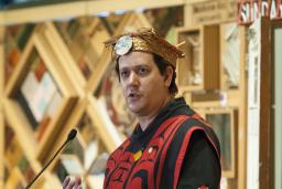 Un homme vêtu d’un costume et d’une coiffe traditionnels autochtones se tient devant une installation d’art faite de panneaux de bois avec des objets encastrés.