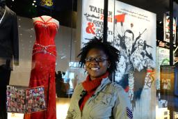 Une femme souriante se tient devant une robe de bal rouge et un smoking noir exposés sur des mannequins. La robe et le costume sont derrière une vitrine.