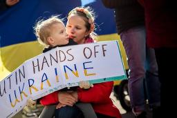 Un enfant tient un panneau de protestation qui dit, en anglais : « Poutine, ne touche pas à l’Ukraine. » L’enfant est assis sur les genoux de la personne qui s'occupe de lui. Derrière eux se trouve un drapeau ukrainien.