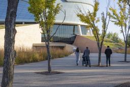 Quatre personnes suivent le chemin menant au Musée canadien pour les droits de la personne par une journée d'automne. Trois d'entre elles sont à pied et une personne est en fauteuil roulant.