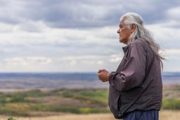 Une personne aux longs cheveux gris se tient à l'extérieur, avec un paysage vallonné et herbeux et un ciel nuageux au loin.