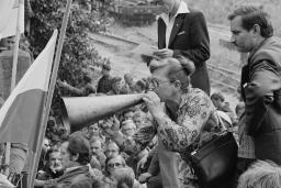 Une femme portant une robe coupe sa main à un mégaphone placé à sa bouche. Elle se tient debout au-dessus d’une foule de personnes rassemblée devant un portail, avec deux drapeaux flottant à proximité.