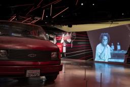 Fourgonnette Chevrolet rouge dans un espace d'exposition avec une vidéo de Rage Against the Machine en arrière-plan.