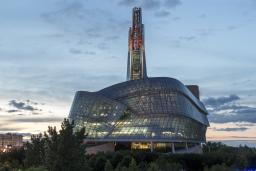 Photo extérieure des fenêtres en verre et de la tour du Musée canadien pour les droits de la personne, prise au crépuscule avec le ciel des prairies en arrière-plan. Le panneau Winnipeg à Forks est situé à droite de l'image. Plusieurs tours de bureaux et les lumières du centre-ville sont visibles à gauche de l'image.