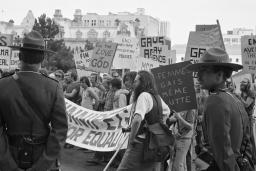 Un grand groupe de protestataires défile dans une ville tandis que deux agents de la GRC en uniforme surveillent la scène. Sur cette image en noir et blanc, les pancartes et les banderoles réclament la fin des pratiques discriminatoires contre les membres de la communauté 2ELGBTQI+.