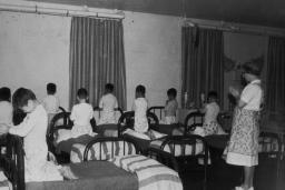 Un groupe de garçons en pyjama est agenouillé sur des lits simples, la tête baissée et les mains jointes comme pour prier. Une femme se tient dans la pièce, les mains jointes de la même manière.