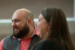 Un homme et une femme sont assis ensemble à une table, souriant chaleureusement tout en écoutant d’autres convives. L’homme porte une chemise saumon et un gilet gris, et de petites bougies et des boissons sont disposées sur la table, créant une atmosphère intime et chaleureuse.