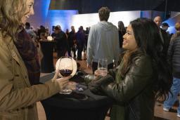 Deux femmes souriantes boivent du vin dans une grande salle remplie de gens.