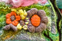 Une œuvre d’art textile colorée qui comprend des fleurs brodées, des branches et de la verdure mélangées à des couches de tissus roses, bleus et blancs.