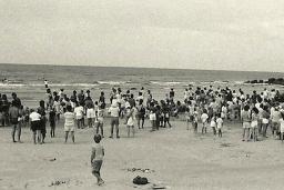 Une photographie en noir et blanc d’une foule de personnes, la plupart debout, sur une plage.